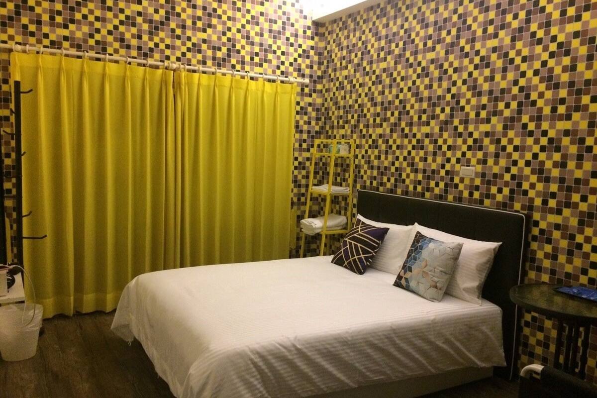 A hotel room with a checkered mosaic wall, a bed with white bedding, a yellow curtain, and a small table with a lamp.
