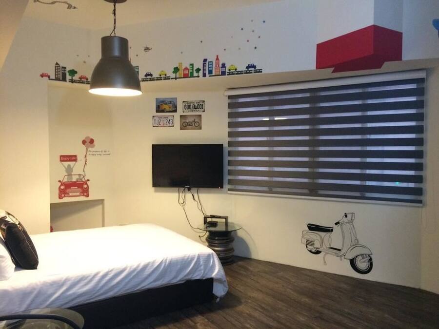 Un dormitorio con cama, televisor montado en la pared, ventana con persianas y un adhesivo decorativo de patinete.