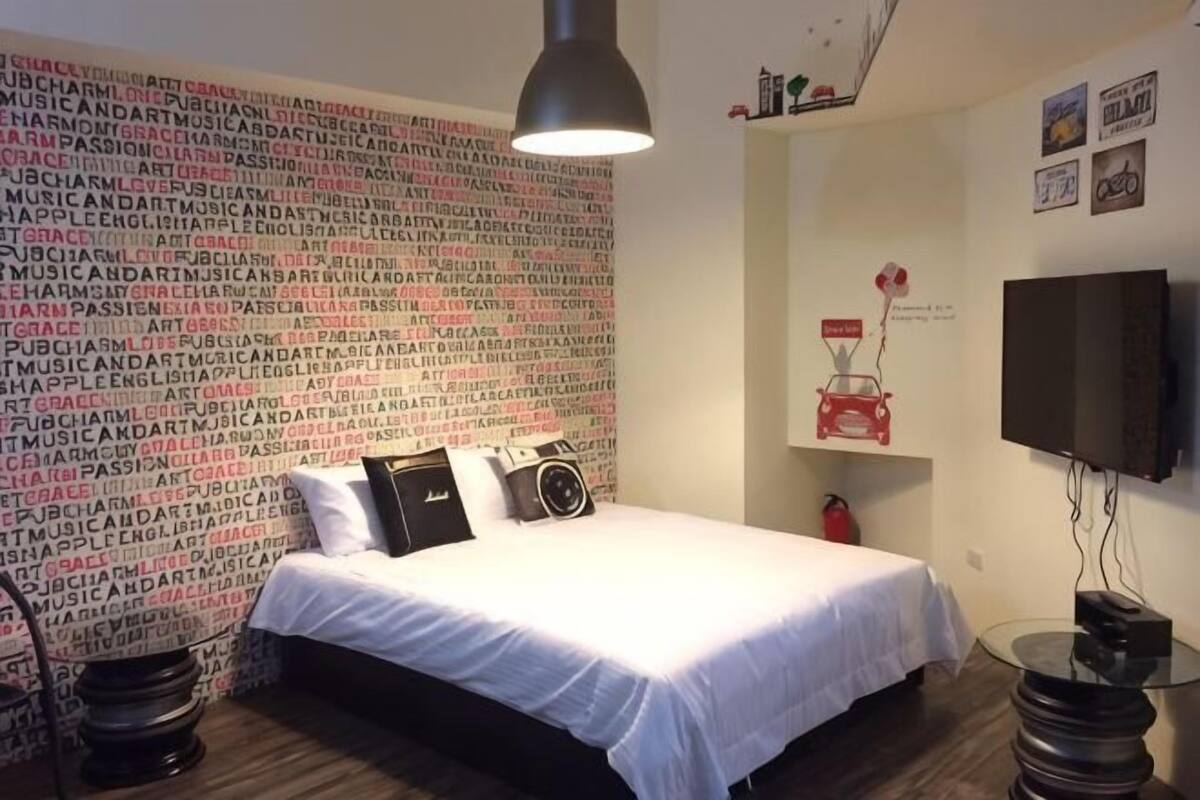 A bedroom with a bed, a wall-mounted TV, a shelf with a red object, and a patterned wall.