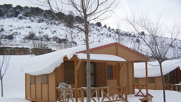 A wooden cabin with a porch and a chair, surrounded by snow. - Camping Los Cameros (Villoslada de Cameros)