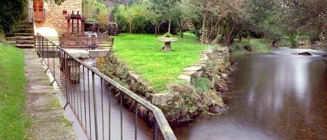 A stone building with a wooden staircase, a grassy area with a stone table, and a calm river.