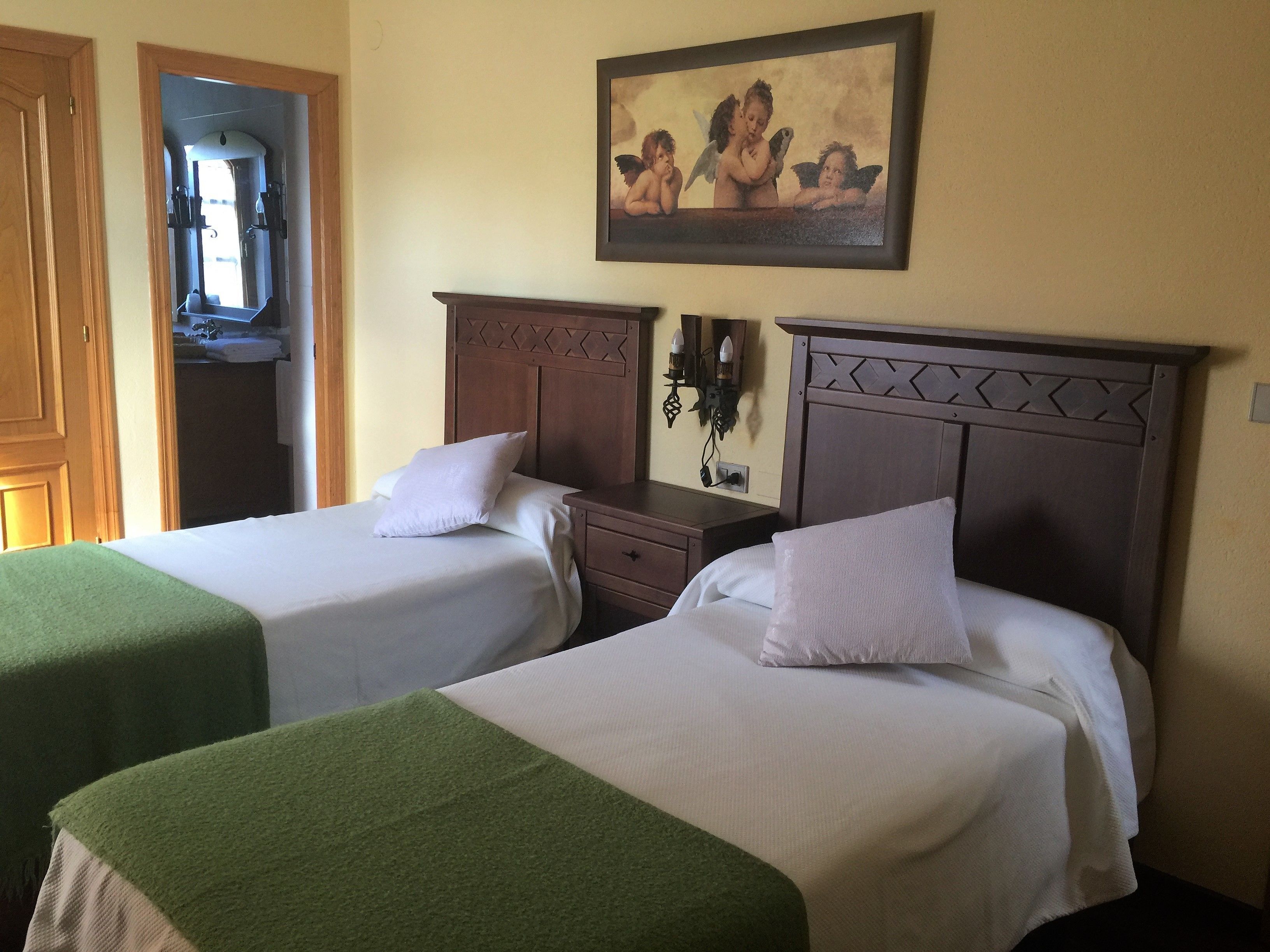 Quarto de hotel com duas camas, cada uma com um travesseiro e um cobertor verde. Há um quadro na parede acima das camas e uma cabeceira de madeira com um padrão decorativo.
