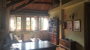 Una habitación rústica con vigas de madera, una gran mesa de madera, un radiador, una estantería y dos cuadros enmarcados en la pared.