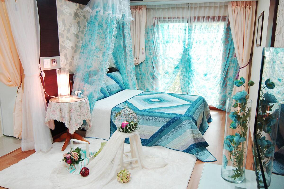 A bedroom with a bed covered in a blue quilt, a bedside table with a lamp, and a vase with flowers.