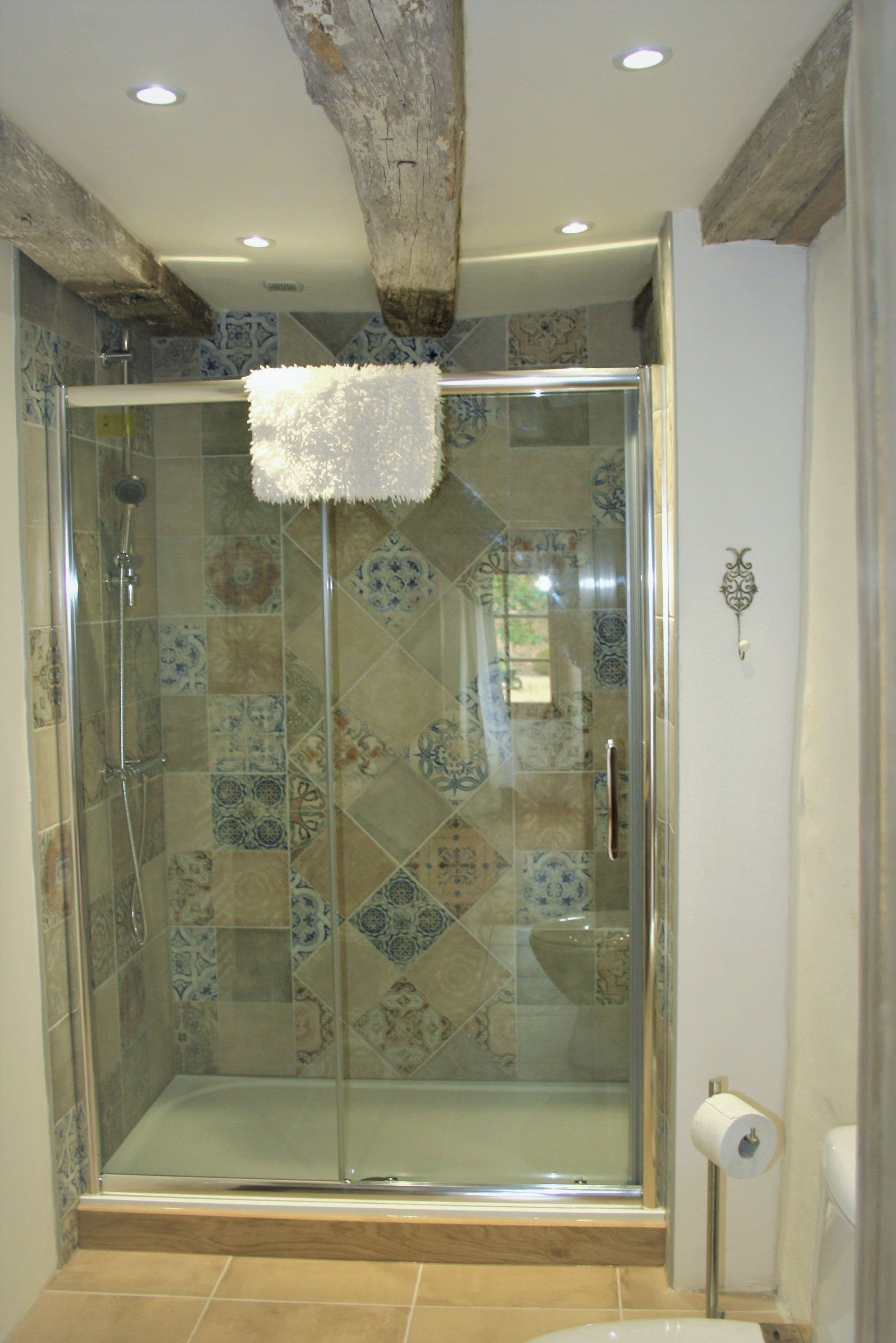 Una cabina doccia in vetro con parete rivestita in piastrelle decorate e trave in legno a soffitto.