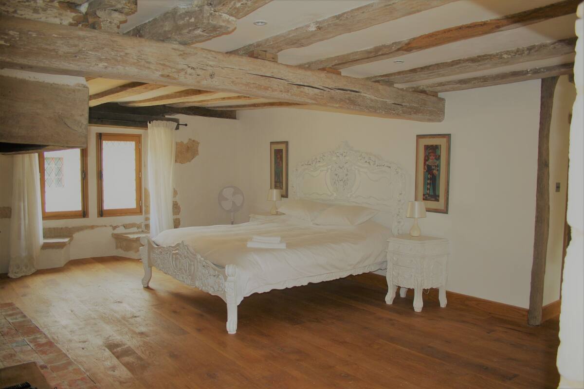 A bedroom with a large bed, wooden beams on the ceiling, a bedside table, and two windows.