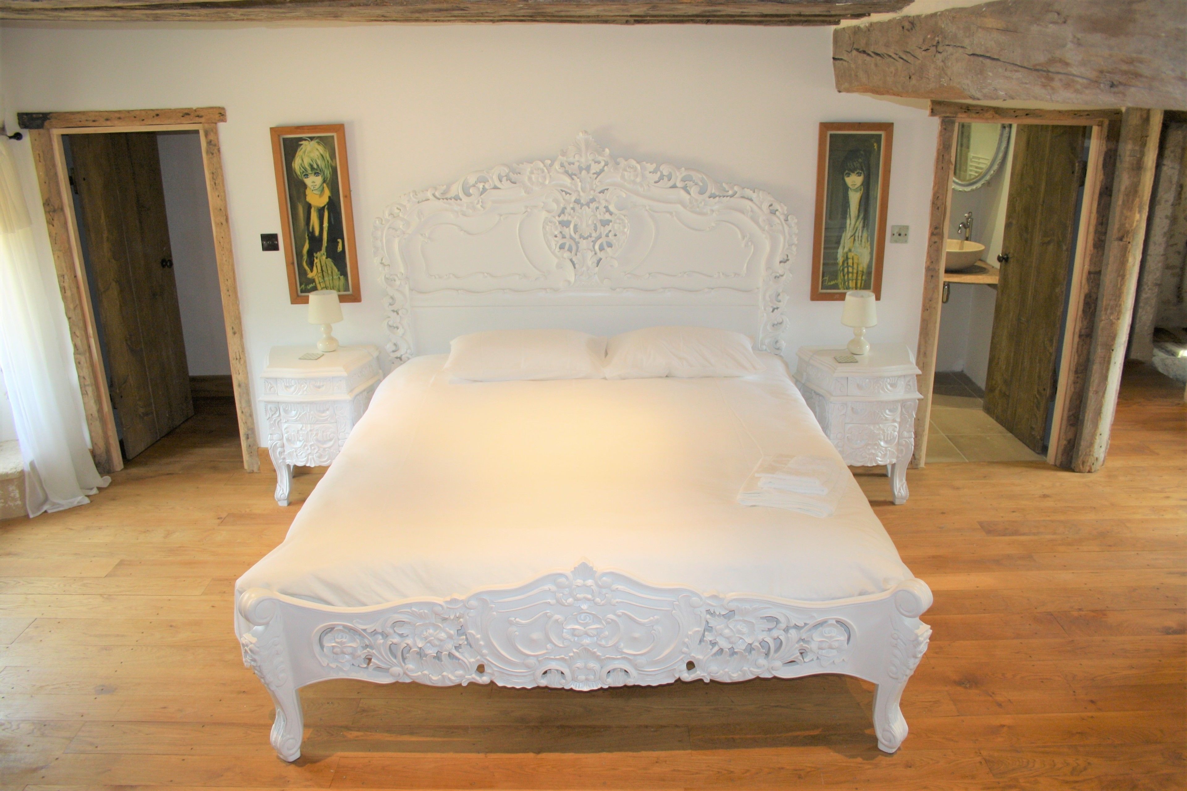 Un letto grande e decorato, con testiera e pediera bianche, affiancato da due comodini con lampade, in una stanza con pavimento in legno e travi a vista.