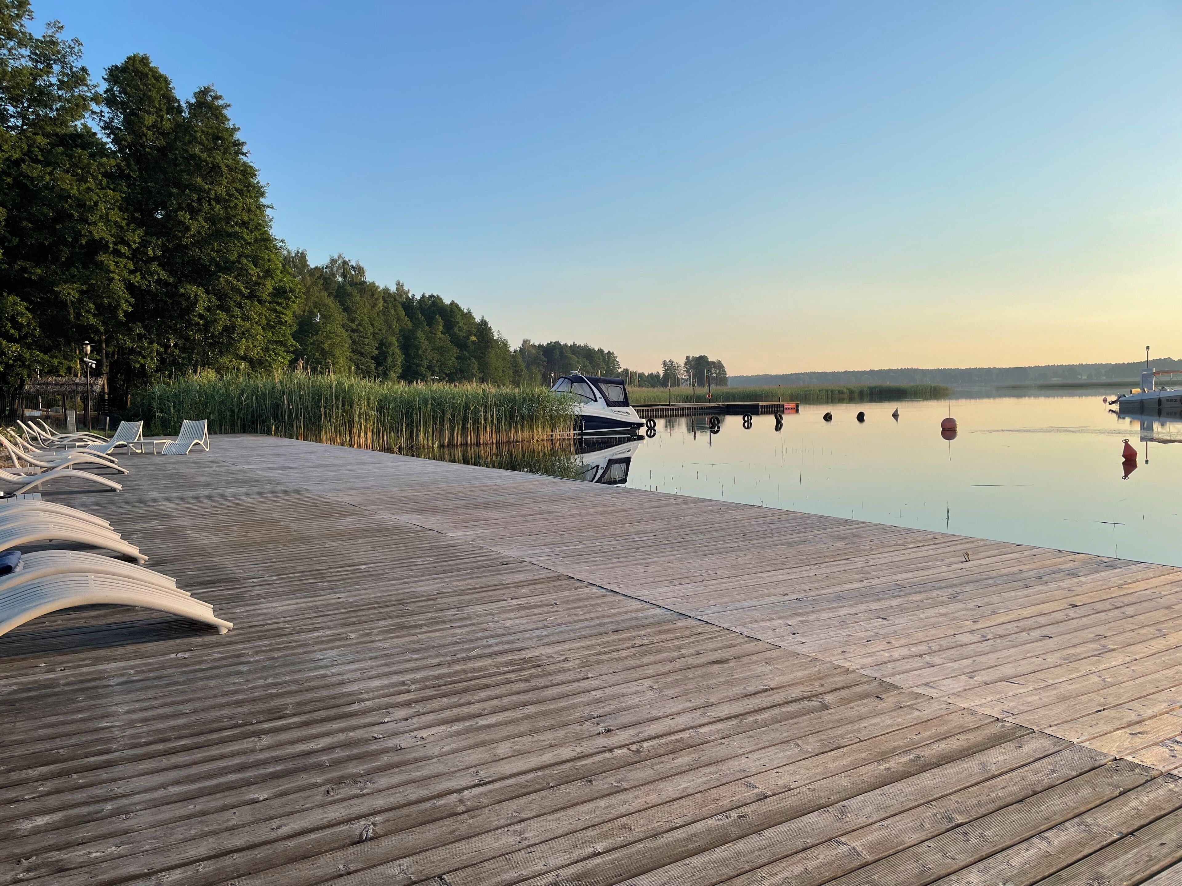 Une jetée en bois avec des chaises longues, un bateau amarré à son extrémité, et un lac paisible, avec des arbres en arrière-plan.