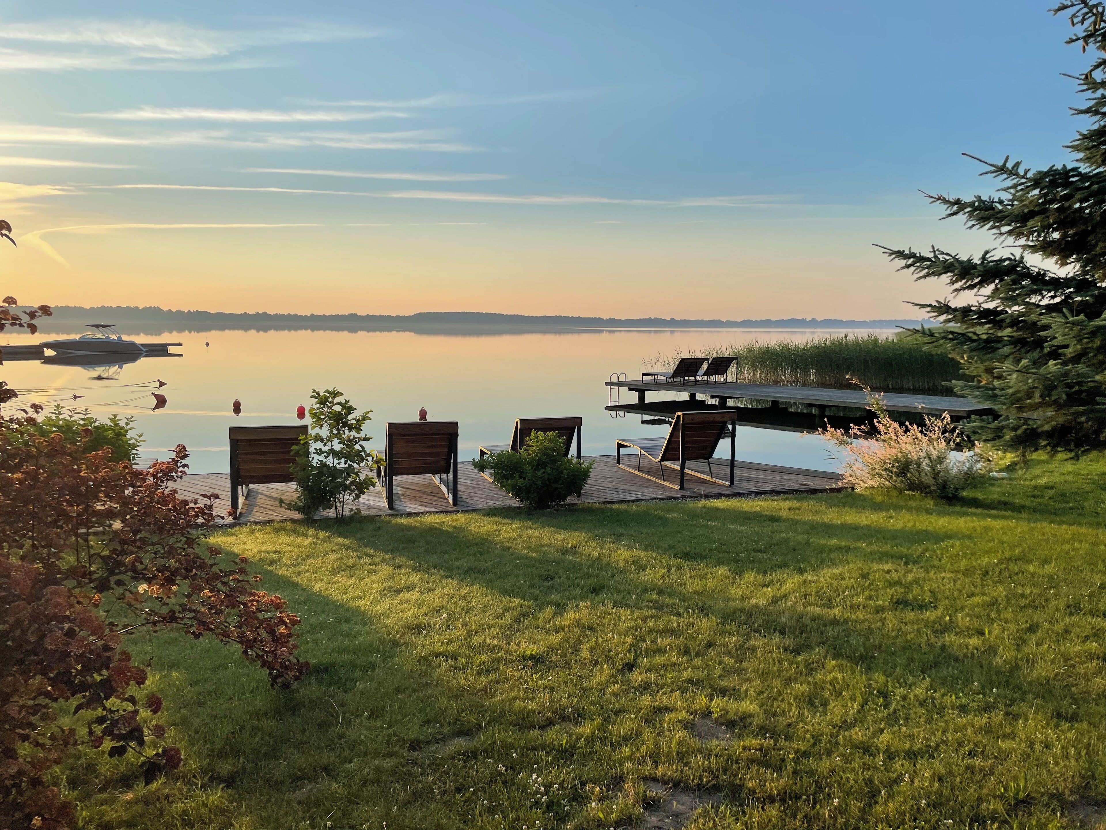 A lakeside deck with wooden chairs and a small table, overlooking a calm lake with a distant shoreline.