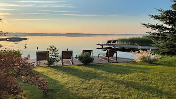 A lakeside deck with wooden chairs and a small table, overlooking a calm lake with a distant shoreline.