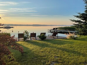 A lakeside deck with wooden chairs and a small table, overlooking a calm lake with a distant shoreline.