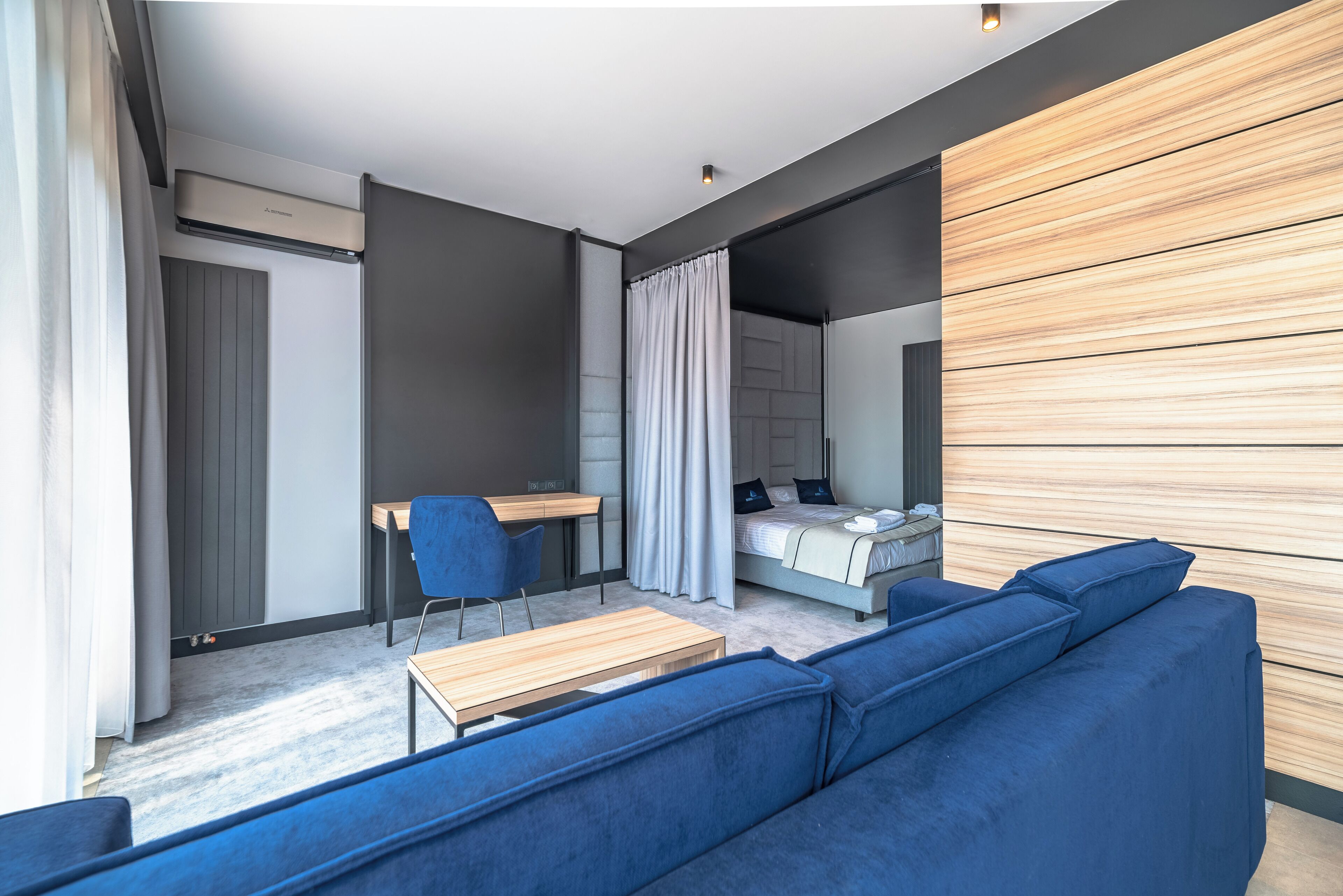 A modern room with a blue sofa, a wooden desk, a bed with a white headboard, and a large window with curtains.