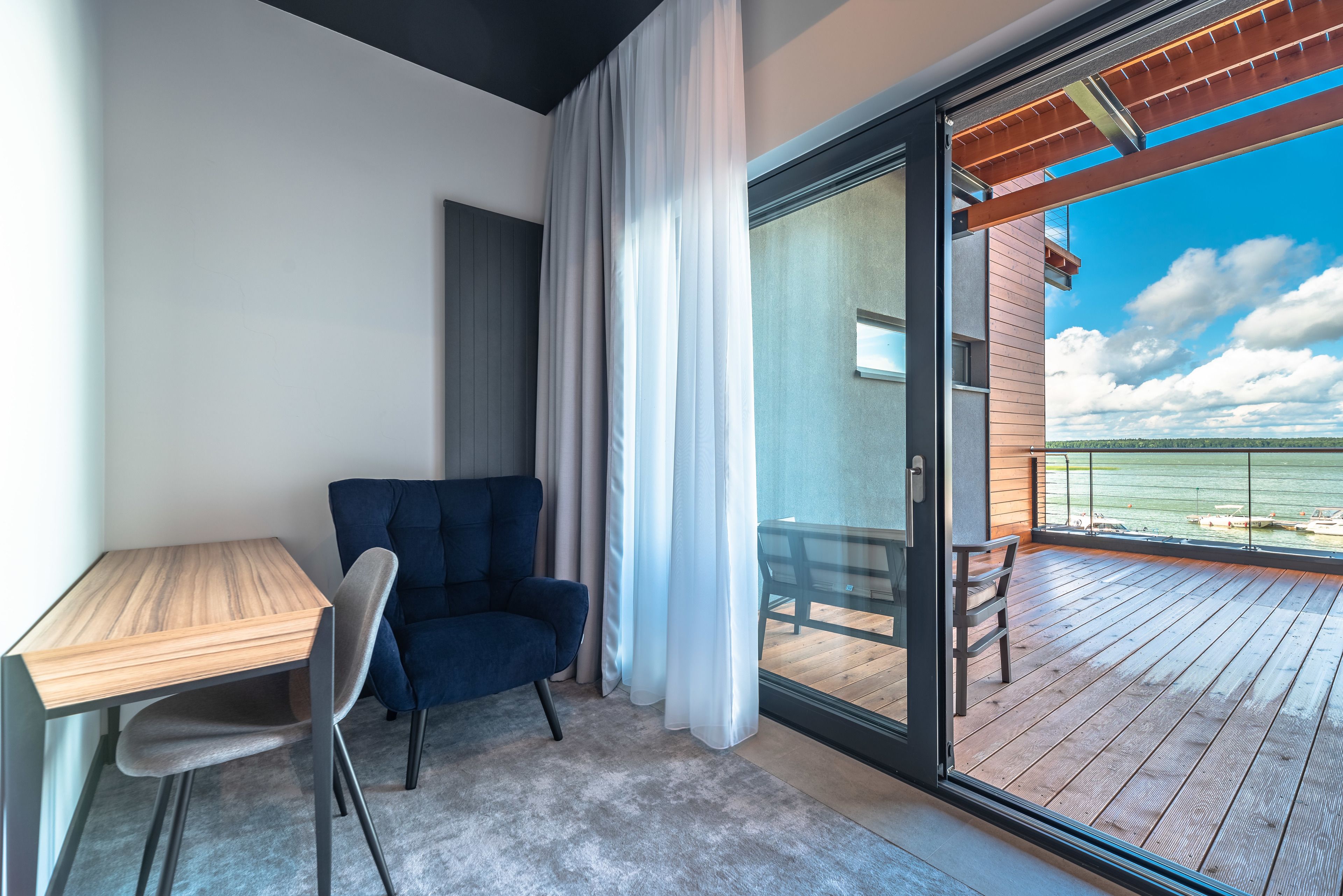Ein modernes Zimmer mit einem Holztisch, einem blauen Stuhl und einer Schiebeglastür, die auf einen Balkon mit Blick auf das Wasser führt.