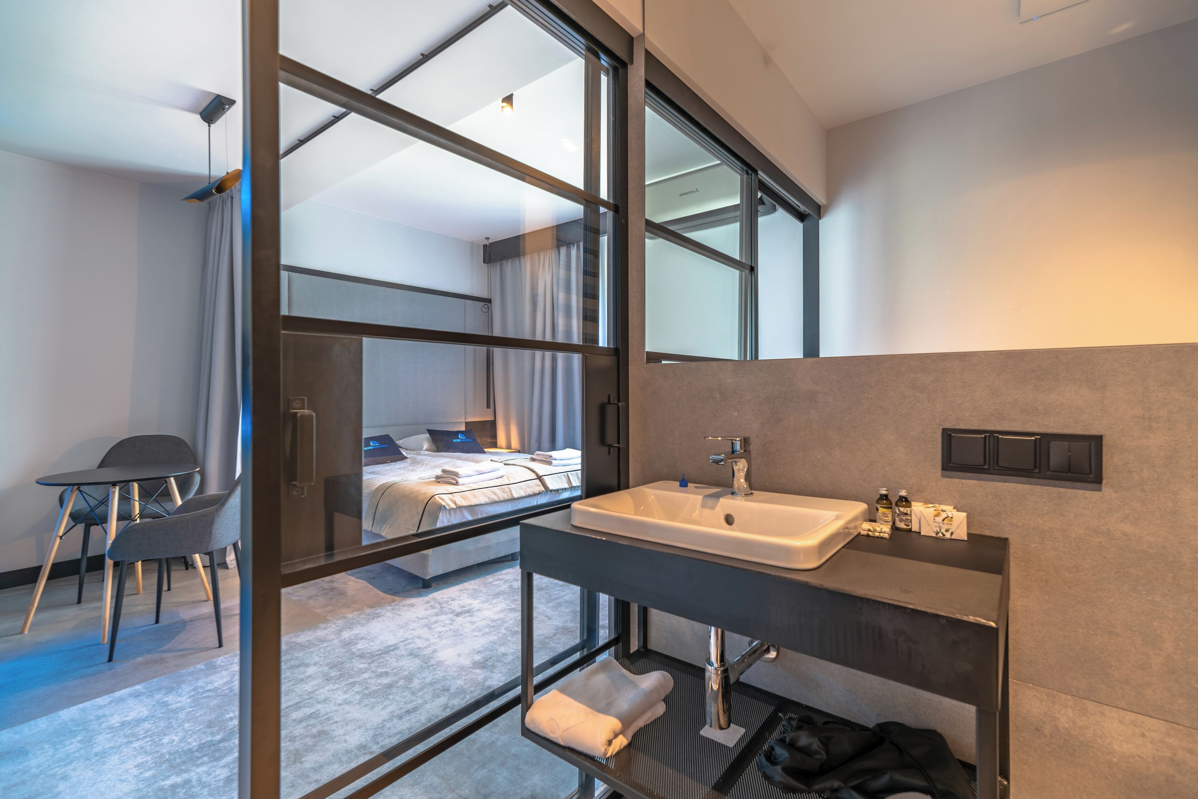 Una habitación de hotel moderna con un ventanal grande, una cama, un escritorio y un baño con lavabo y espejo.
