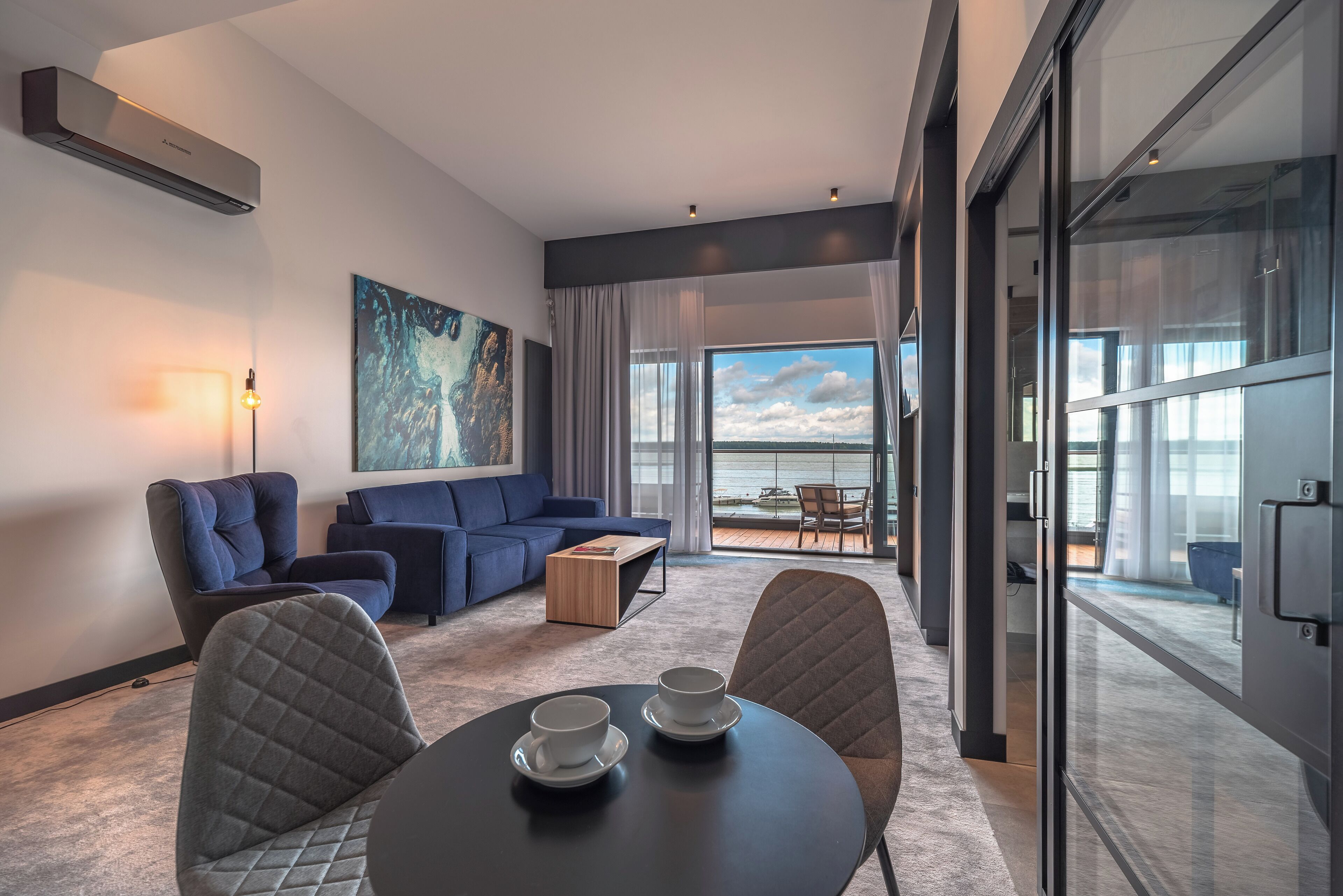 A modern hotel room with a large window, a sofa, a coffee table, and a dining table with two cups.