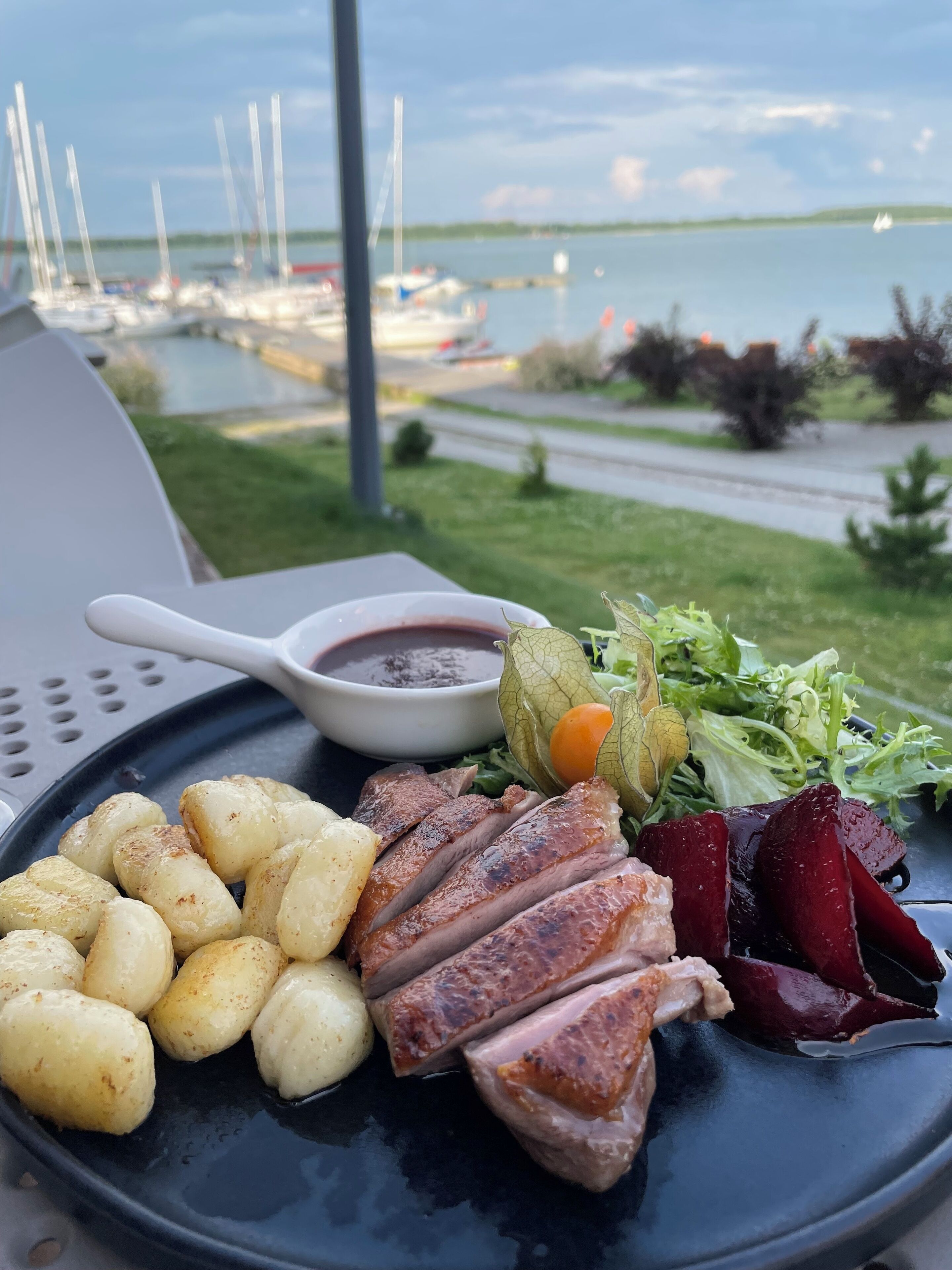 A plate of food with roasted potatoes, duck, beets, and greens, with a view of a marina in the background.