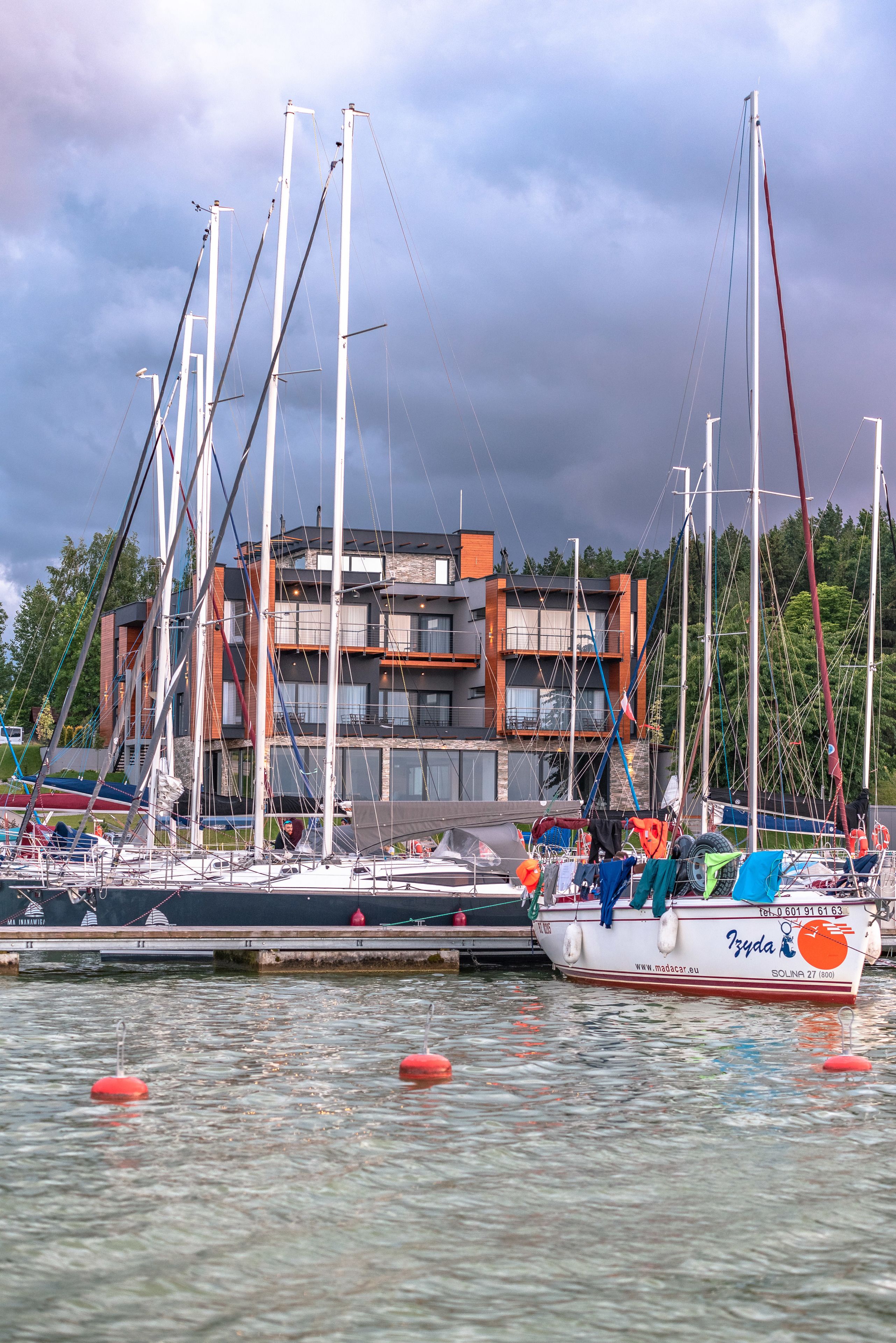A marina with several sailboats docked, a modern building with balconies, and a calm water surface.