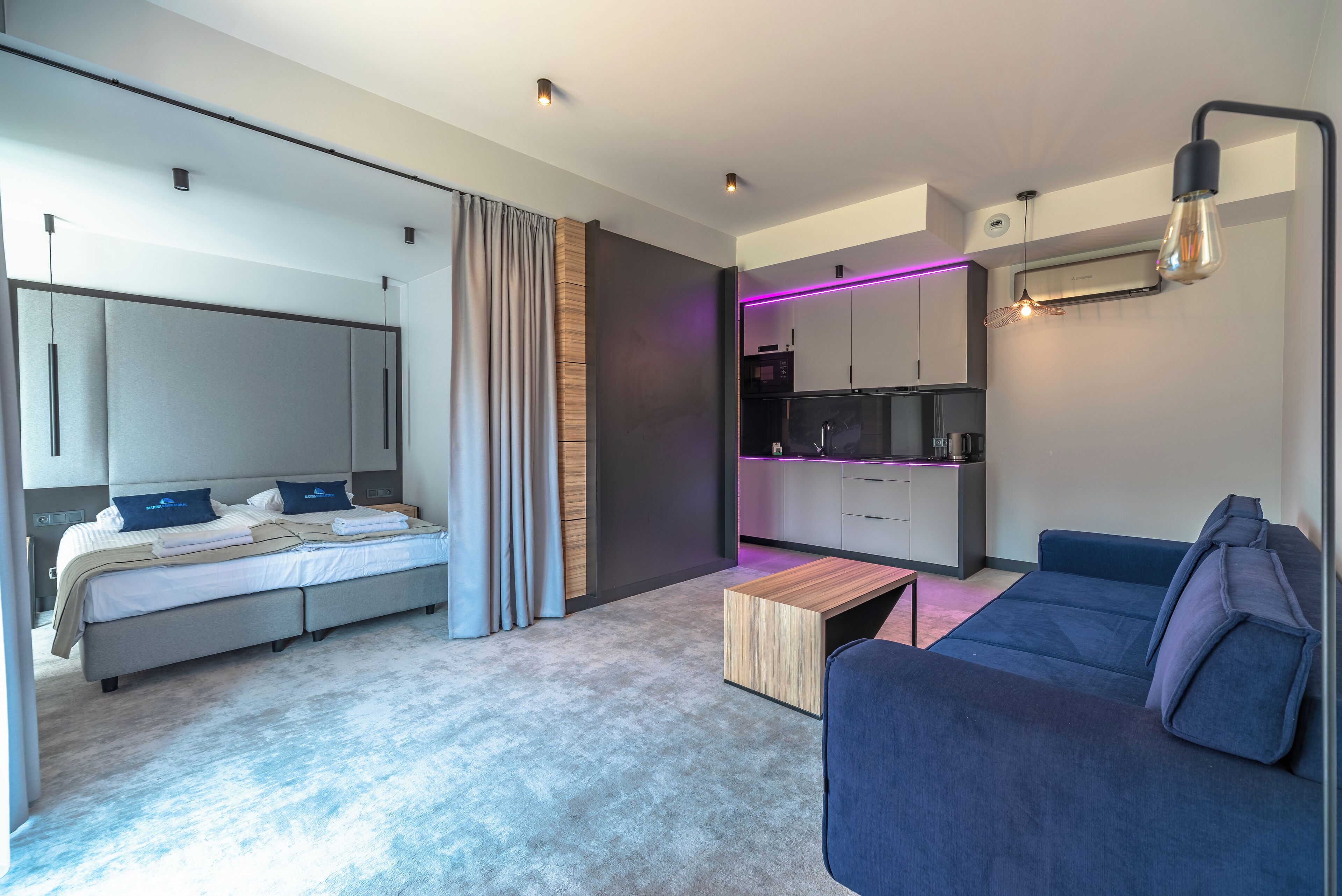 A modern hotel room with a bed, a sofa, a small wooden table, and a kitchenette.