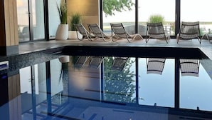 Una piscina interior moderna con paredes de vidrio transparente, sillones de descanso y una lámpara de techo de diseño geométrico.