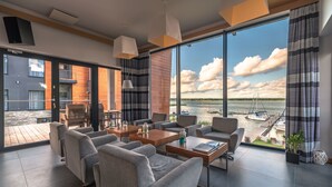 Un salón moderno con ventanas amplias, sofás grises, mesas de madera y vista a un puerto deportivo.