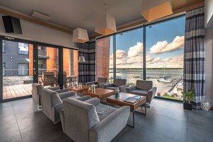 Un salón moderno con ventanas amplias, sofás grises, mesas de madera y vista a un puerto deportivo.