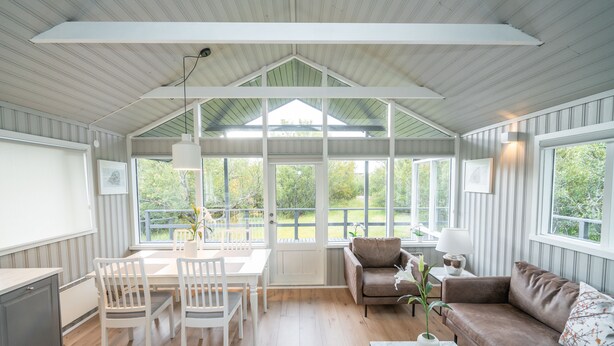 A spacious room with a high ceiling, a dining area with a table and chairs, a brown leather sofa, and a large window offering a view of the outdoors. - Húsafell Holiday Homes (Borgarbyggd)