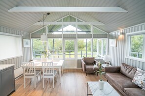 A spacious room with a high ceiling, a dining area with a table and chairs, a brown leather sofa, and a large window offering a view of the outdoors. - Húsafell Holiday Homes (Borgarbyggd)