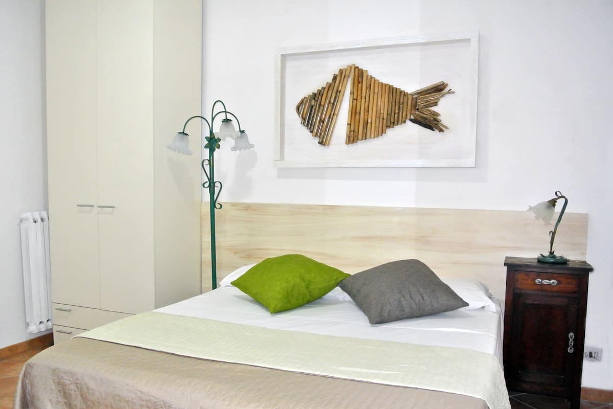 Ein ordentlich bezogenes Bett mit einem grün-grauen Kopfkissen, einer Nachttischlampe und einem gerahmten Fischbild an der Wand.