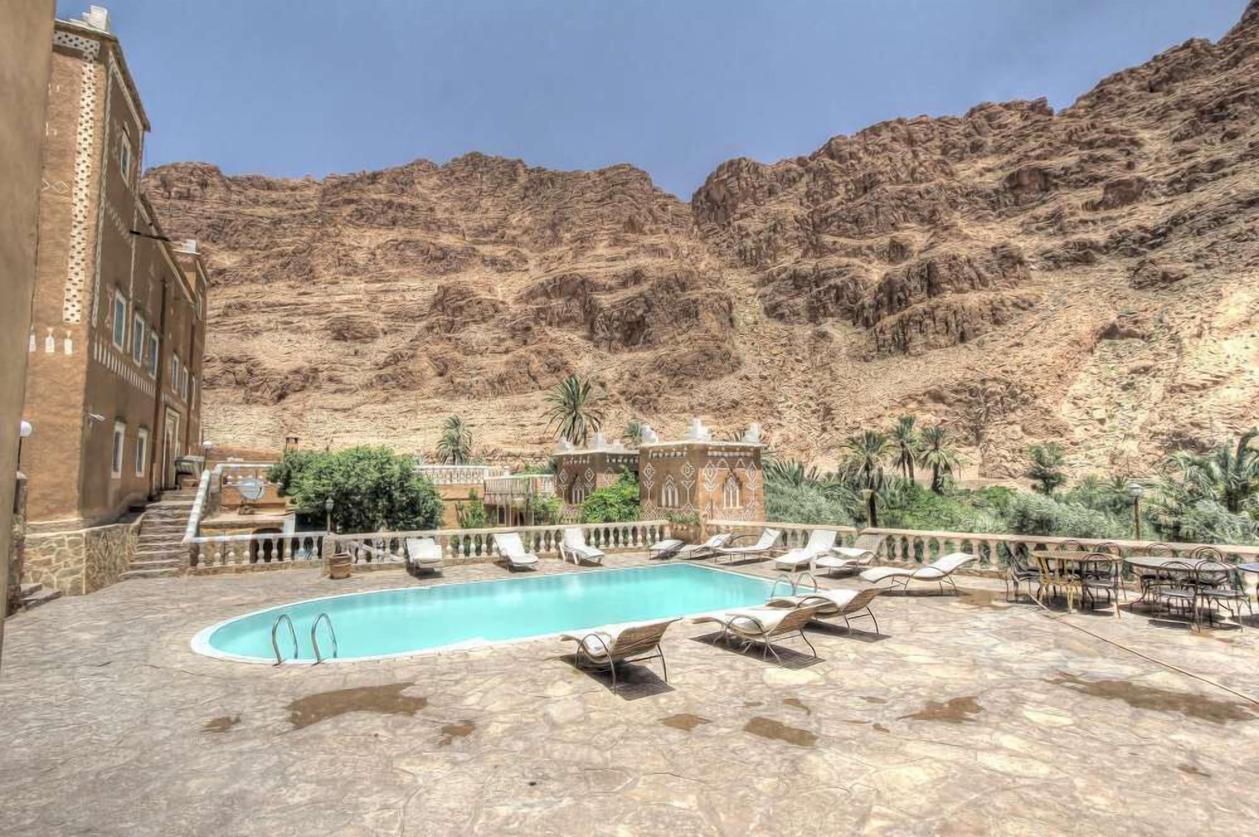 A pool area with lounge chairs and a pool, surrounded by a building with traditional architecture, set against a mountainous backdrop.