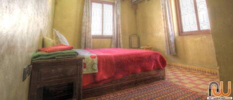 Un dormitorio con una cama de madera, una mesita de noche pequeña, una alfombra estampada y un ventanal con cortinas.