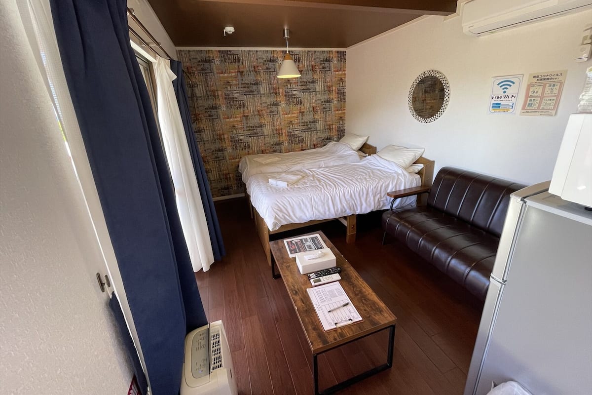 Une chambre d’hôtel de taille modeste, comprenant un lit, un petit canapé, une table en bois avec des papiers, un radiateur fixé au mur et un ventilateur de plafond.