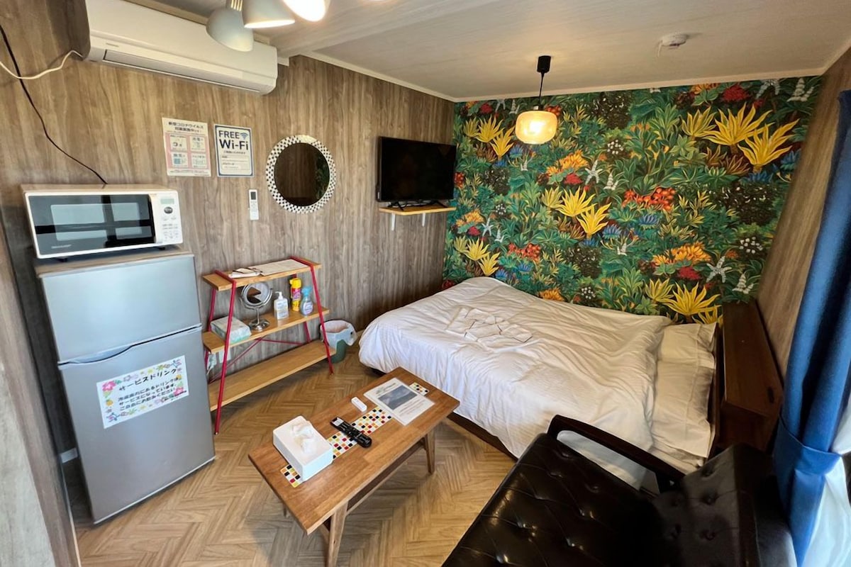 A compact hotel room with a bed, a desk, a microwave, a refrigerator, a TV, and a wall with a floral mural.