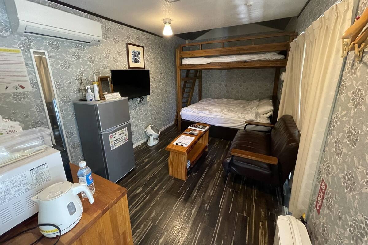 Une chambre compacte comprenant un lit superposé, un petit bureau avec une bouilloire, un réfrigérateur et un téléviseur.