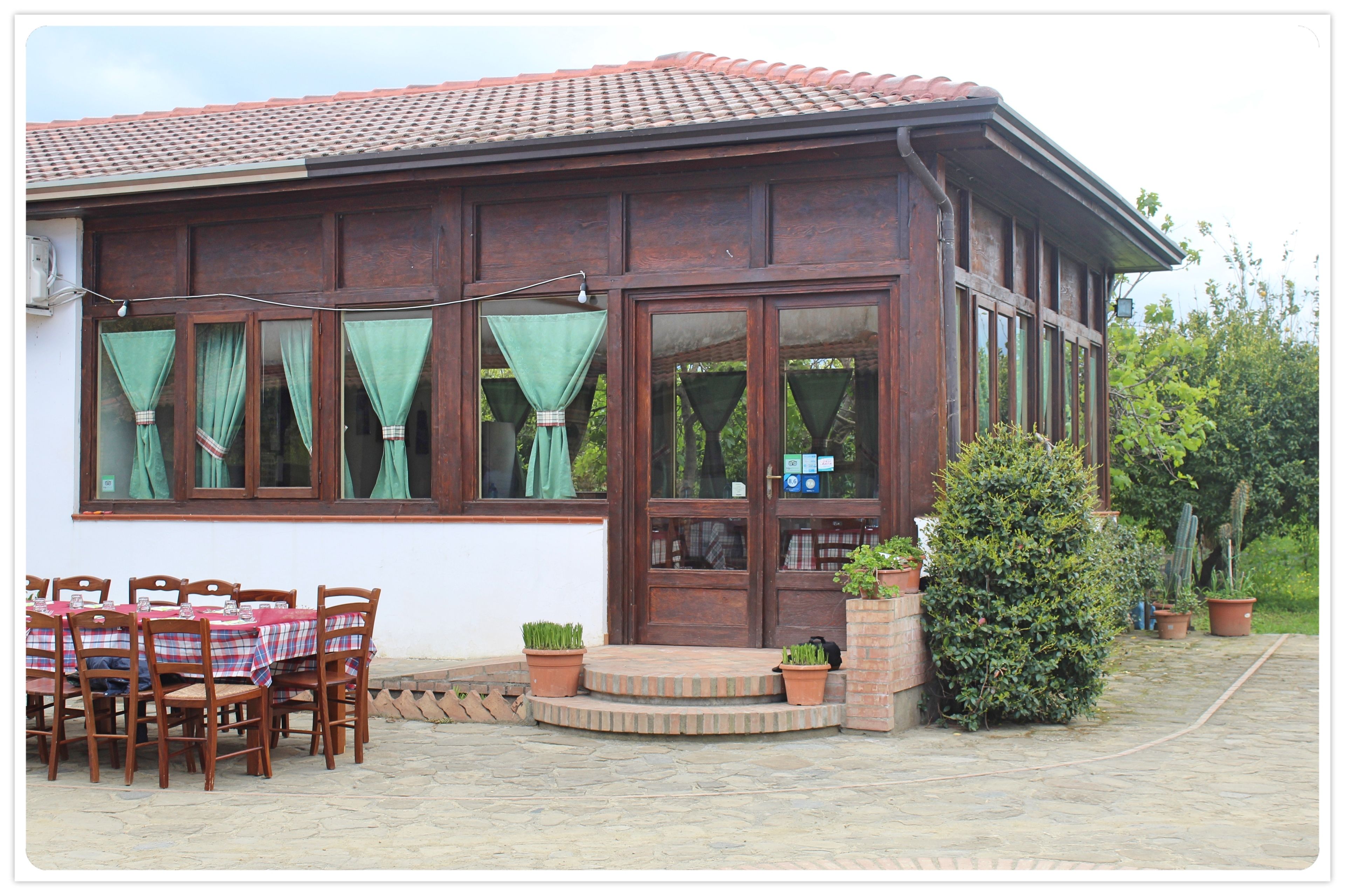 A wooden building with a tiled roof, large windows with curtains, a brick entrance, and a patio with tables and chairs.