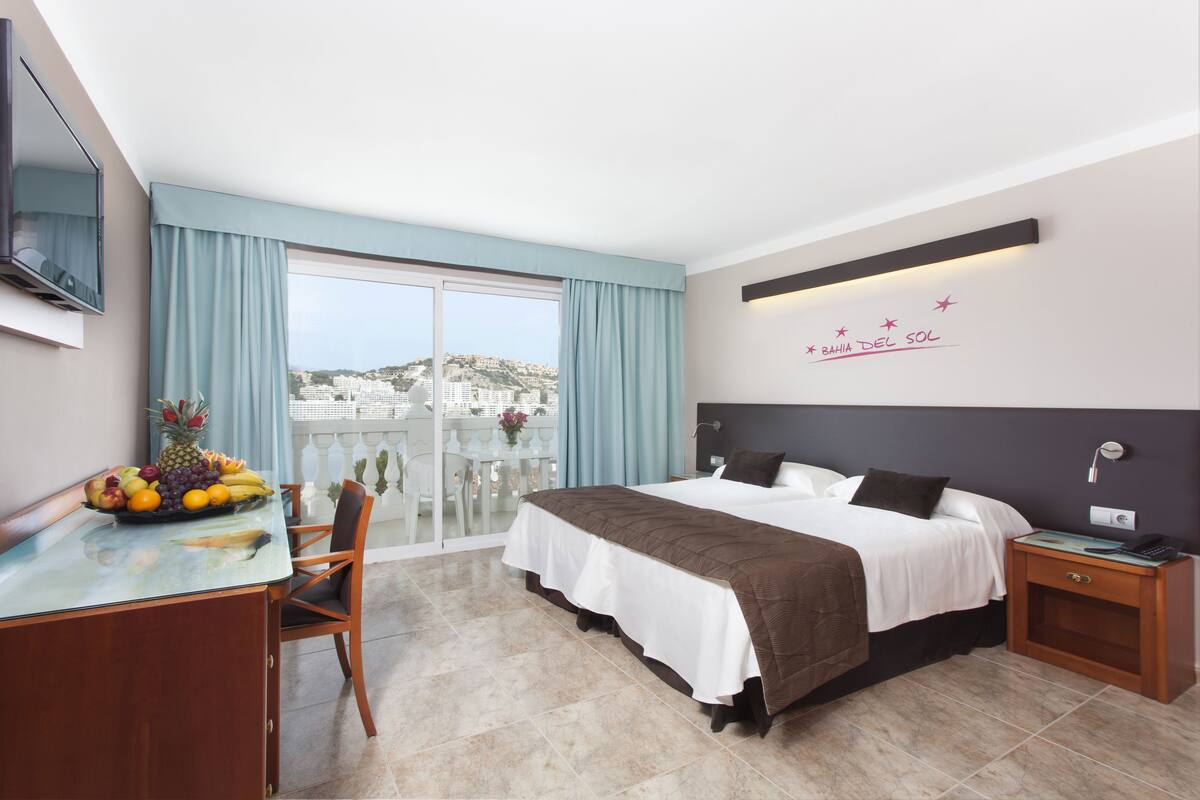 Quarto de hotel com uma cama, uma escrivaninha com uma cesta de frutas, uma cadeira, uma mesa de cabeceira e vista para a paisagem urbana.