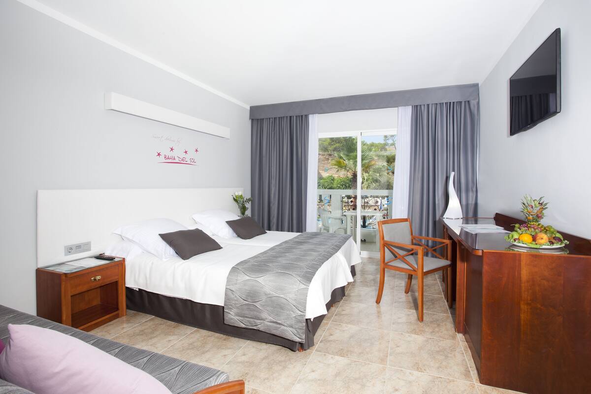 Quarto de hotel com cama, escrivaninha, cadeira, mesa pequena com frutas e varanda com cortinas.