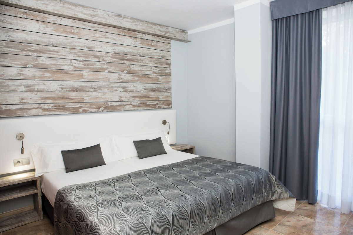 Un dormitorio moderno con cabecera de madera, una cama grande con cubrecama estampado, mesitas de noche con lámparas y vistas a una ventana con cortinas.
