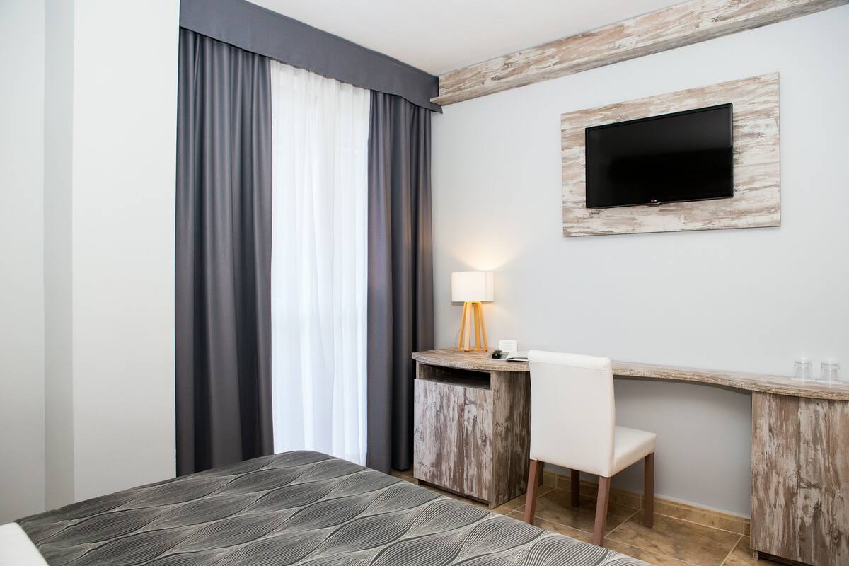 Quarto de hotel com cama, escrivaninha e cadeira, televisão na parede e janela com cortinas.