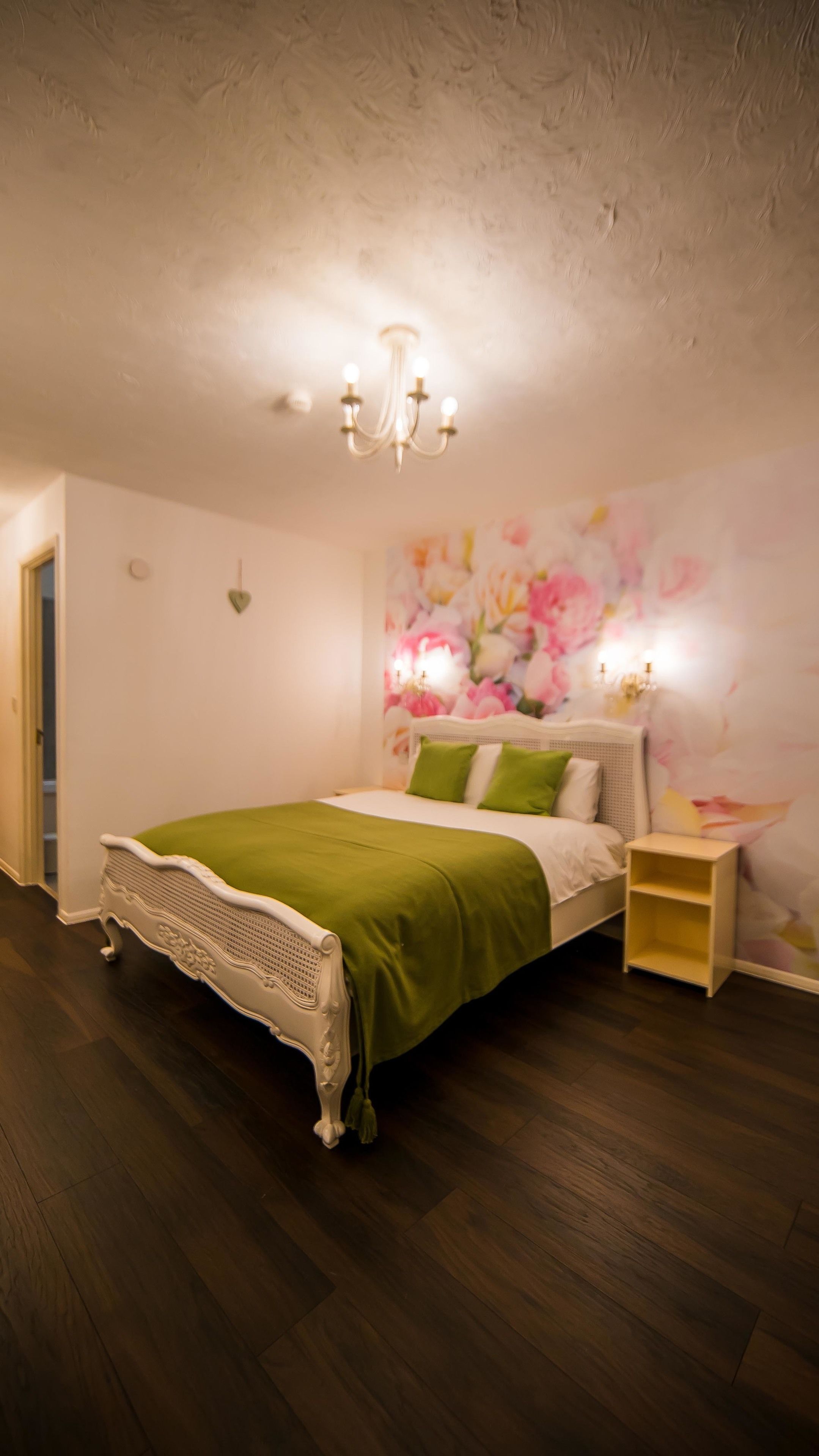 Une chambre avec un plancher en bois, un lit blanc avec une literie verte, une table de chevet et une fresque murale à motifs floraux.