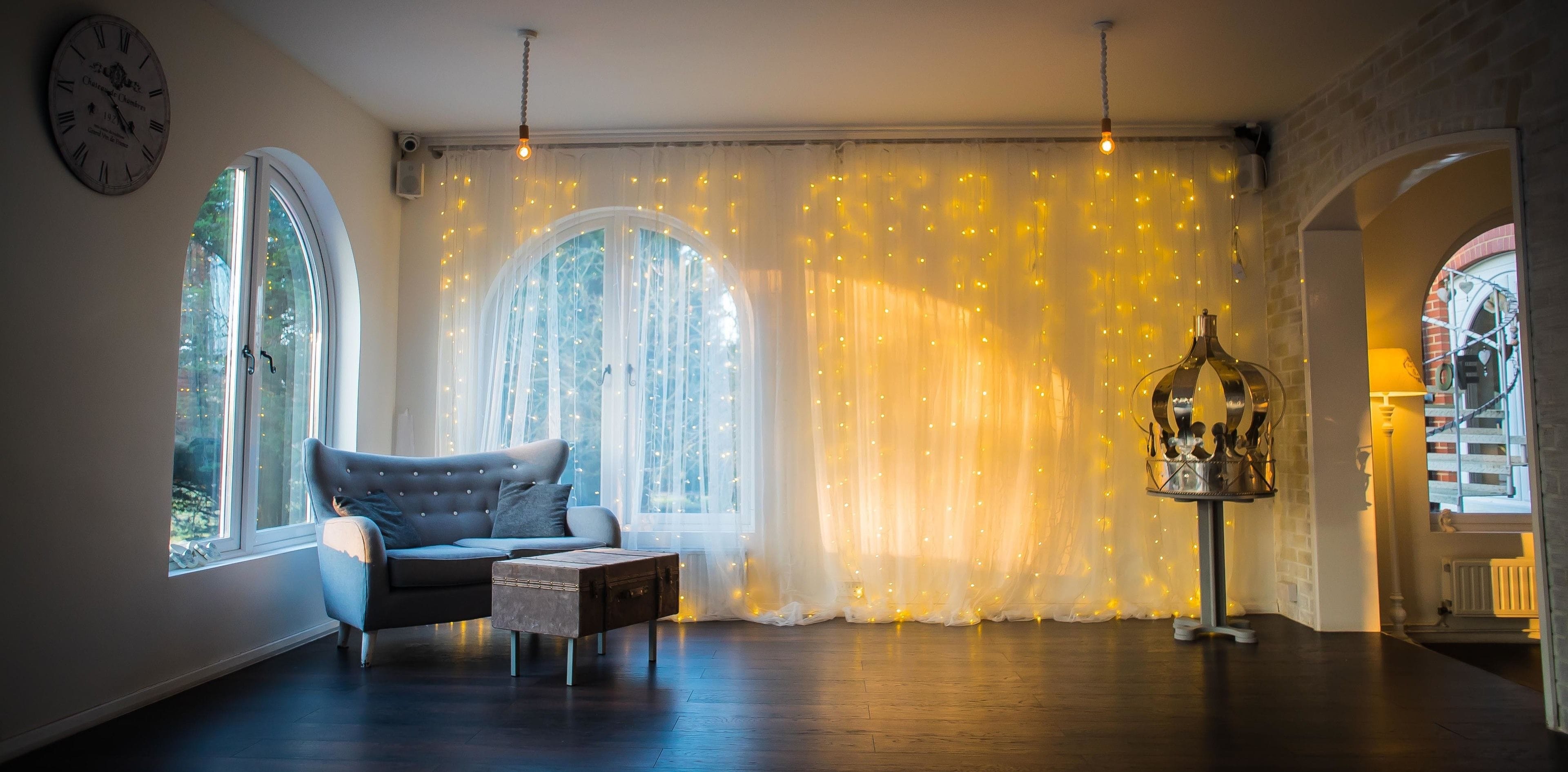 A cozy room with a grey sofa, a small wooden table, and a decorative birdcage. There are two large windows with sheer curtains and fairy lights, and a clock on the wall.