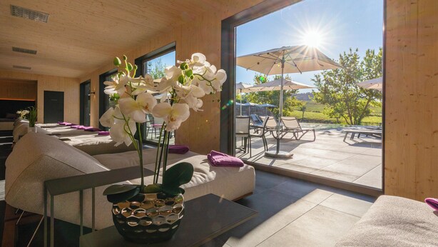 A modern interior with a sofa, a table with a vase of flowers, and a view of an outdoor patio with umbrellas and chairs. - FAIR RESORT All Inclusive Wellness & SPA Hotel Jena (Zollnitz)