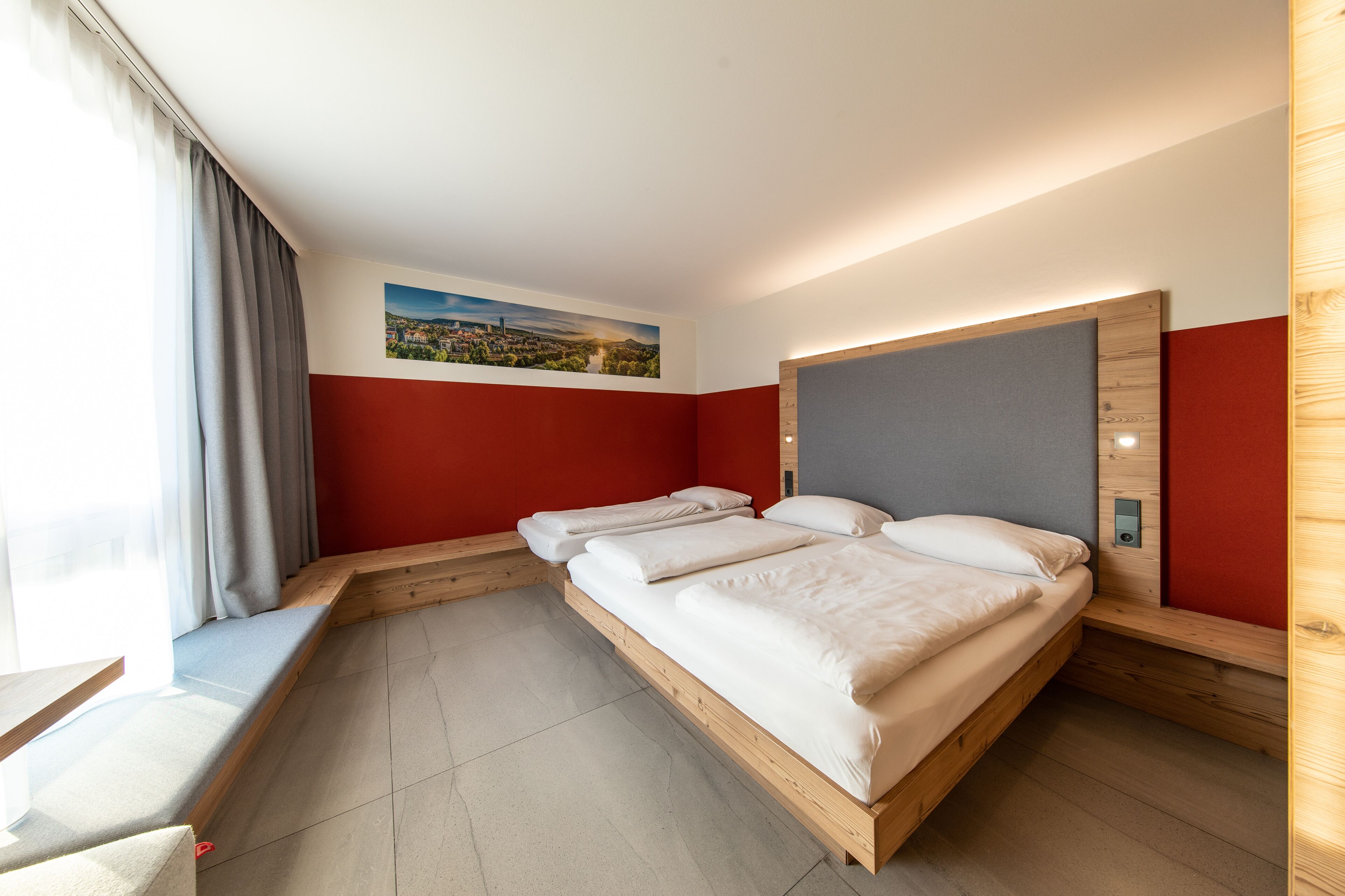 Ein modernes Hotelzimmer mit zwei Einzelbetten, einer Sitzbank am Fenster und einem Stadtbild an der Wand.