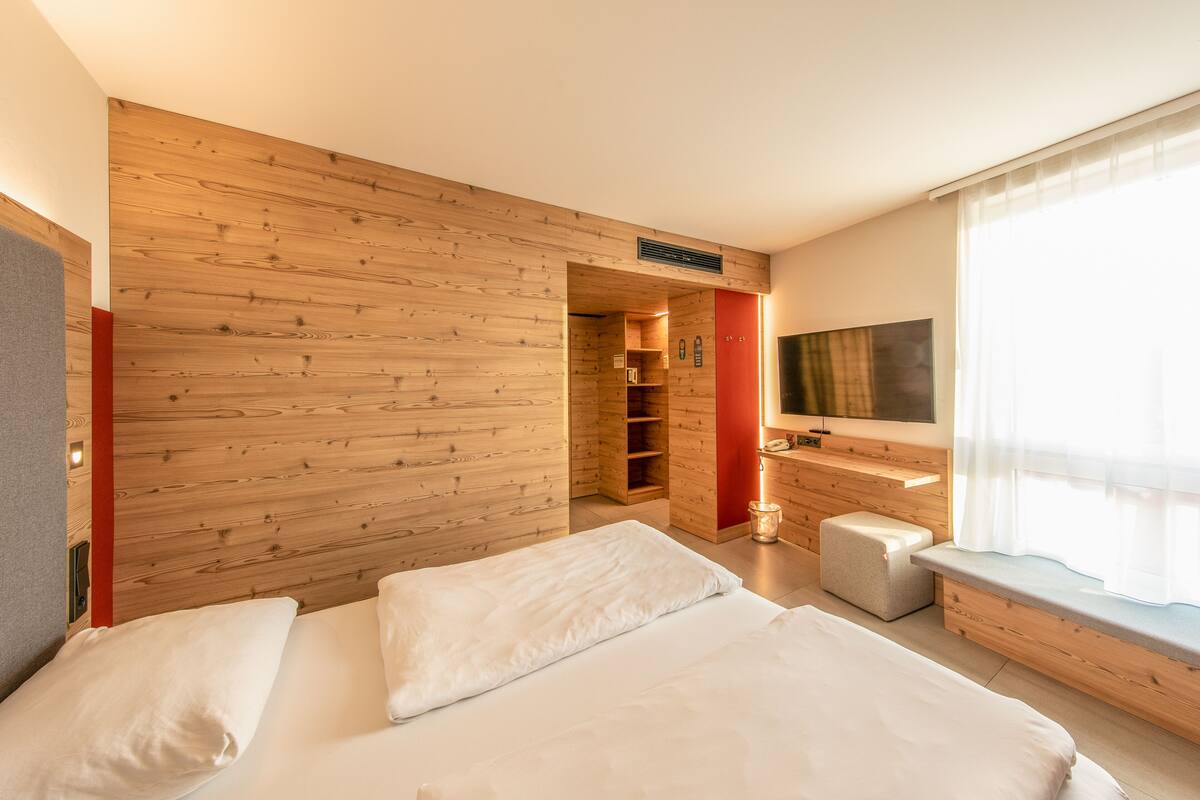 Une chambre d’hôtel moderne avec un mur en bois, un lit avec du linge de lit blanc, un petit coin salon et un téléviseur fixé au mur.