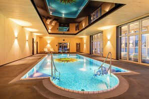 An indoor swimming pool with a sun design on the floor, wooden walls, and a view of the outside through large windows. - FAIR RESORT All Inclusive Wellness & SPA Hotel Jena (Zollnitz)
