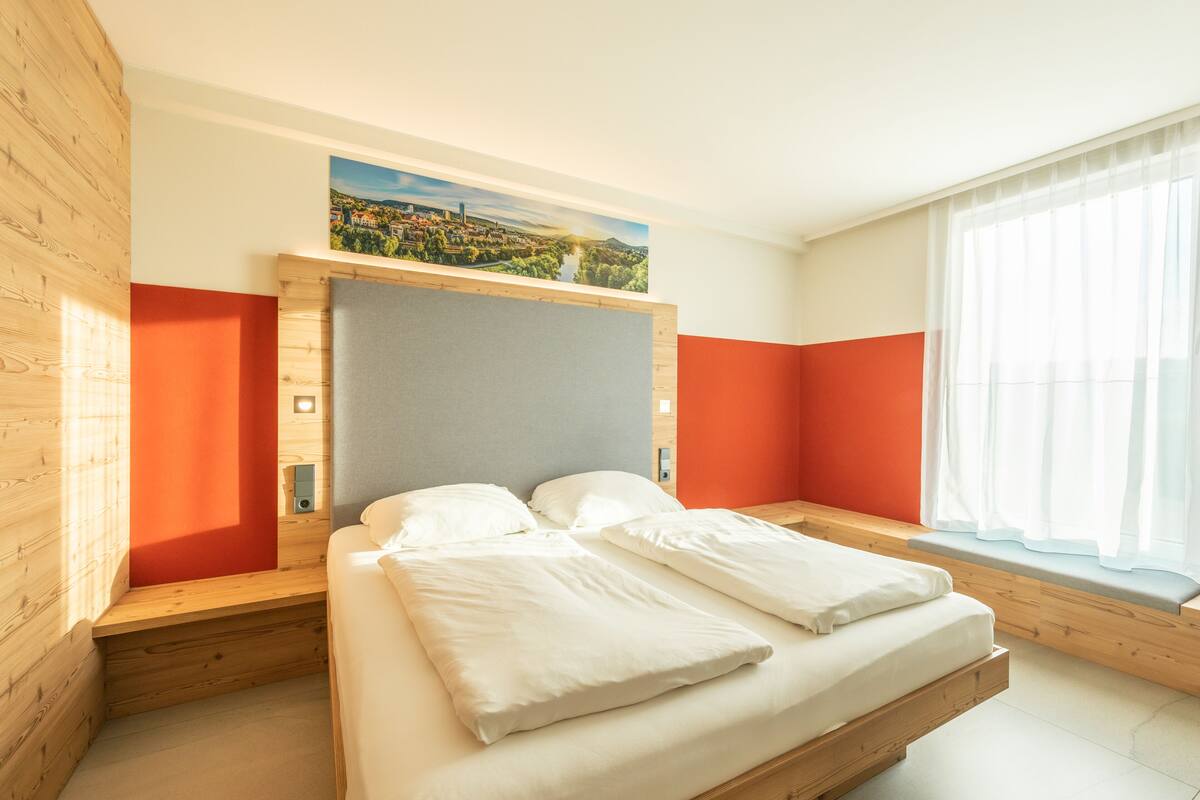 Une chambre moderne avec un lit, un lambris en bois, un tableau représentant un paysage urbain et une fenêtre avec des rideaux.