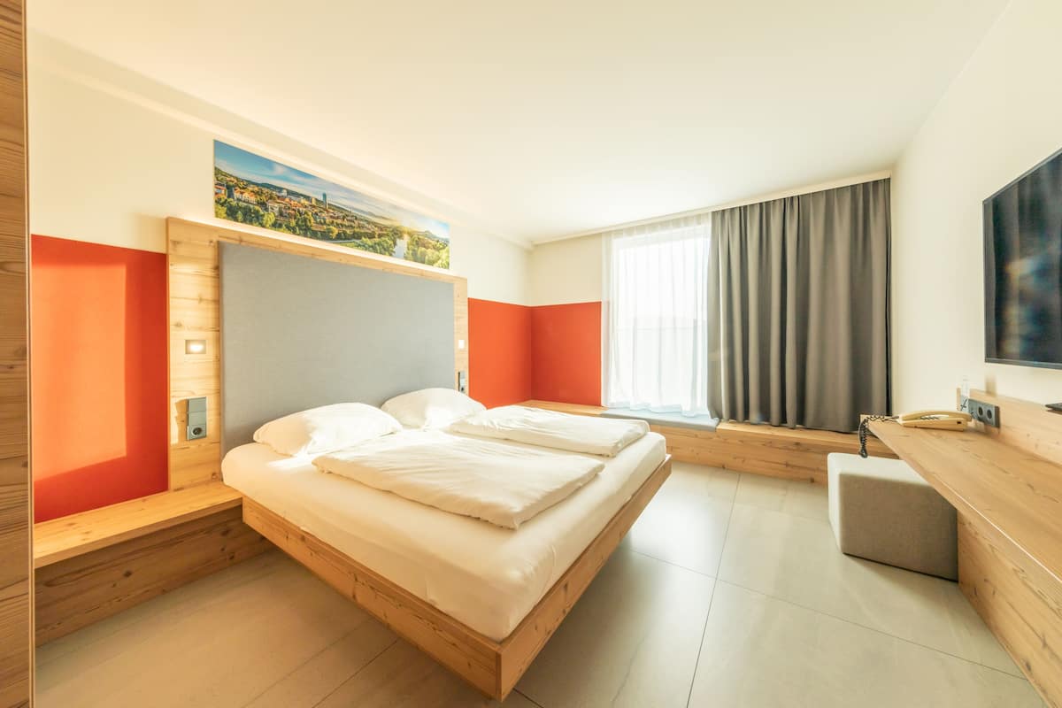 Une chambre d’hôtel moderne avec un lit en bois, un bureau et une grande fenêtre avec des rideaux.