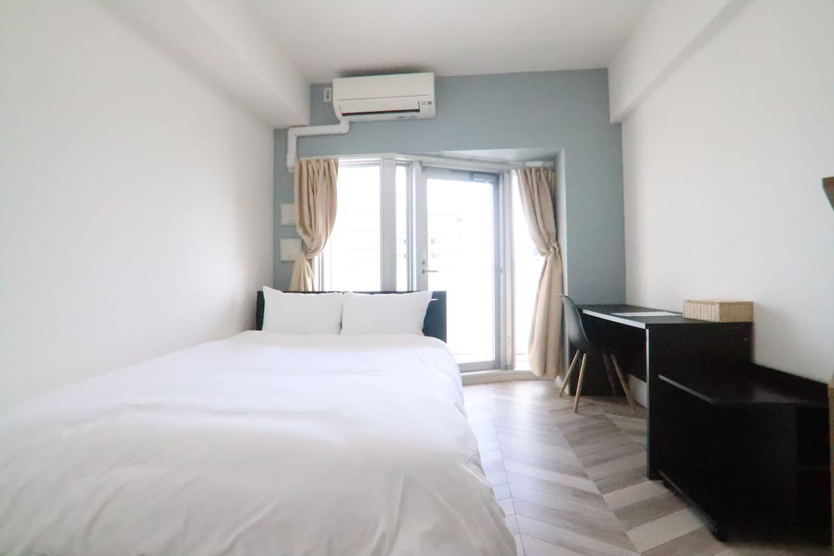 A hotel room with a bed, a desk, and a chair. There is a window with curtains, and an air conditioning unit on the wall.
