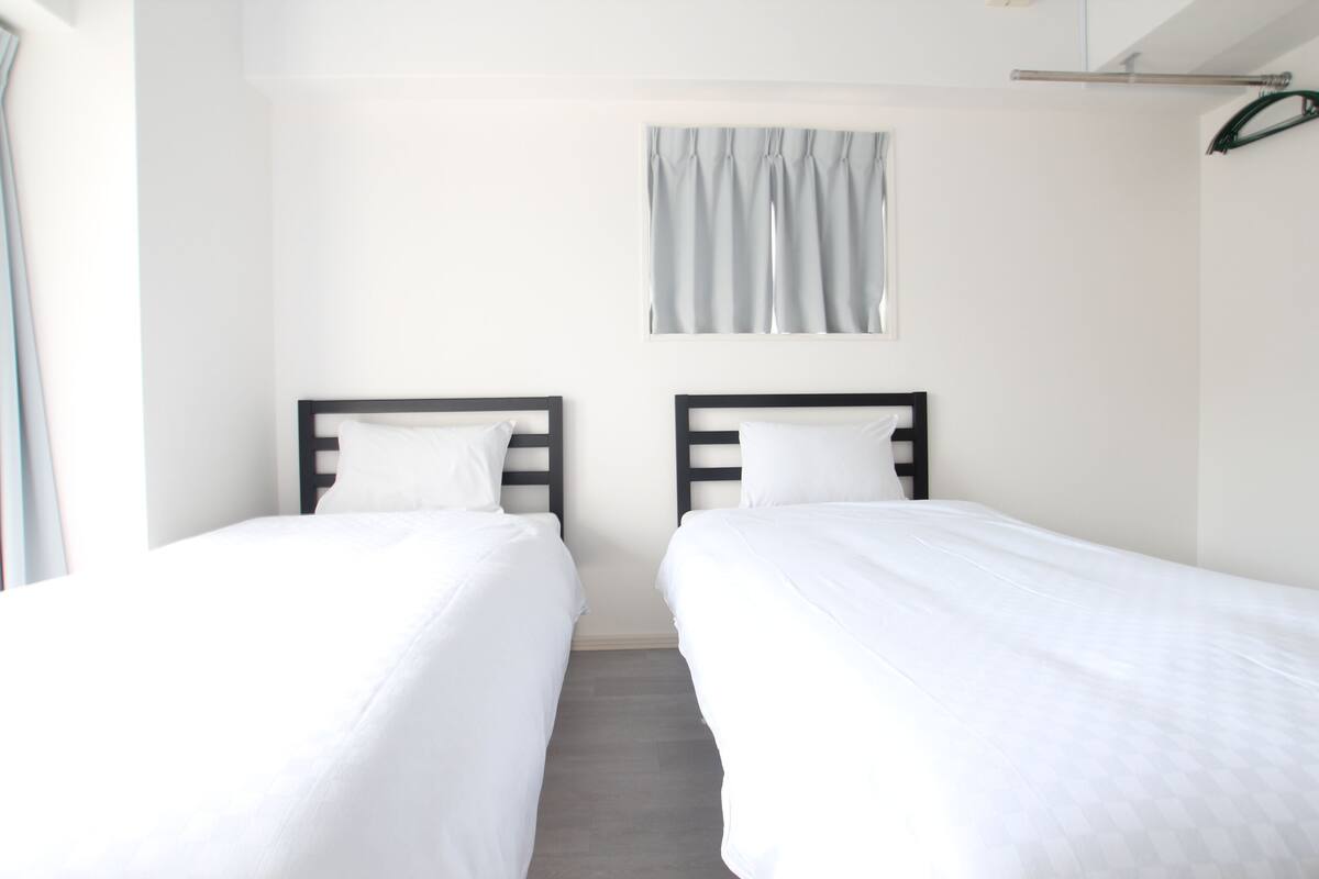 A room with two single beds, white bedding, a window with curtains, and a metal rack on the wall.