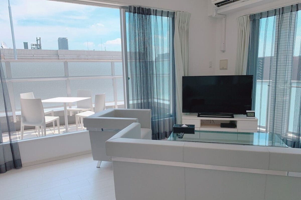 A modern living room with a large TV, white furniture, and a balcony with a city view.