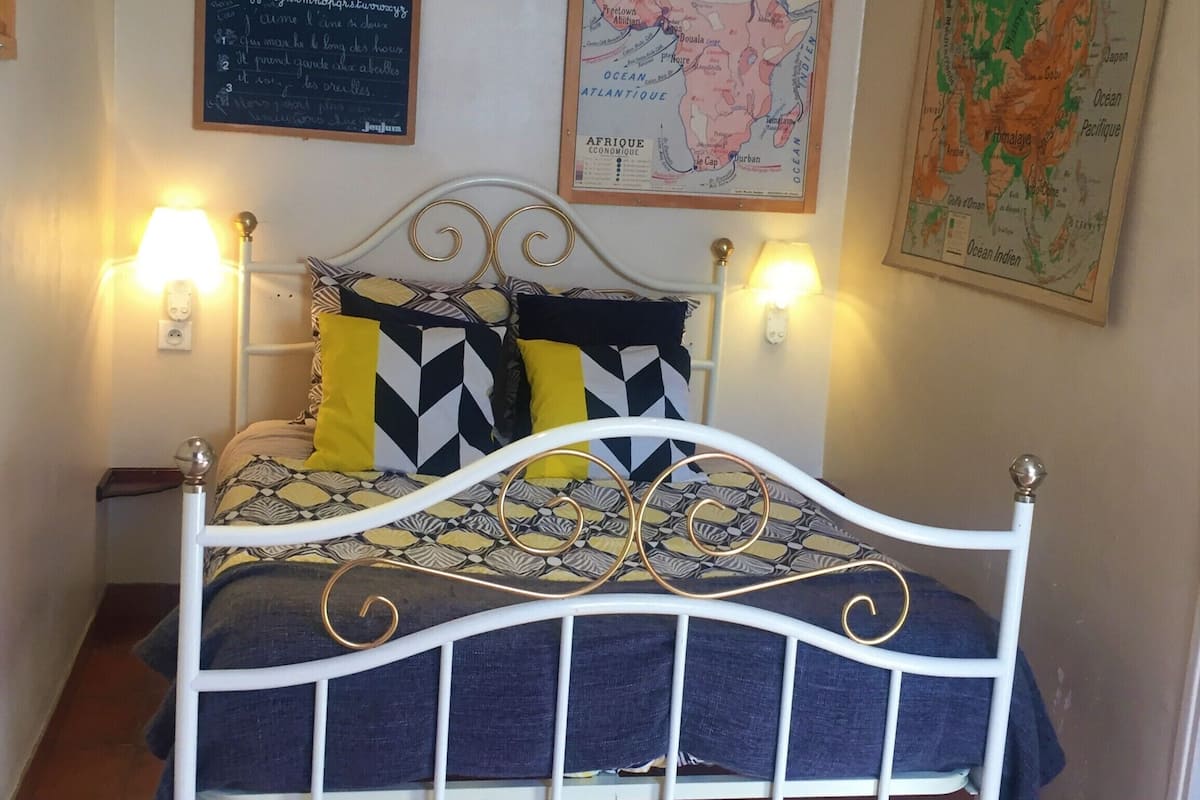 A bed with a white metal frame, a patterned bedspread, and two decorative pillows. There are two wall-mounted lamps on either side of the bed.